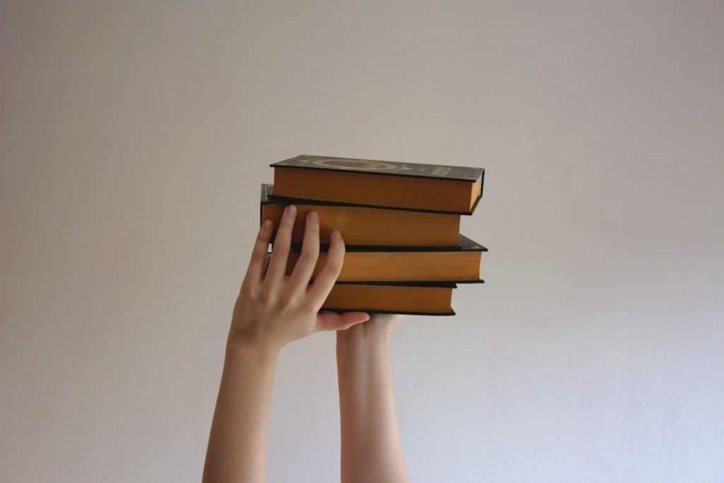 Hands reaching upwards holding a stack of hardbound books against a plain background.