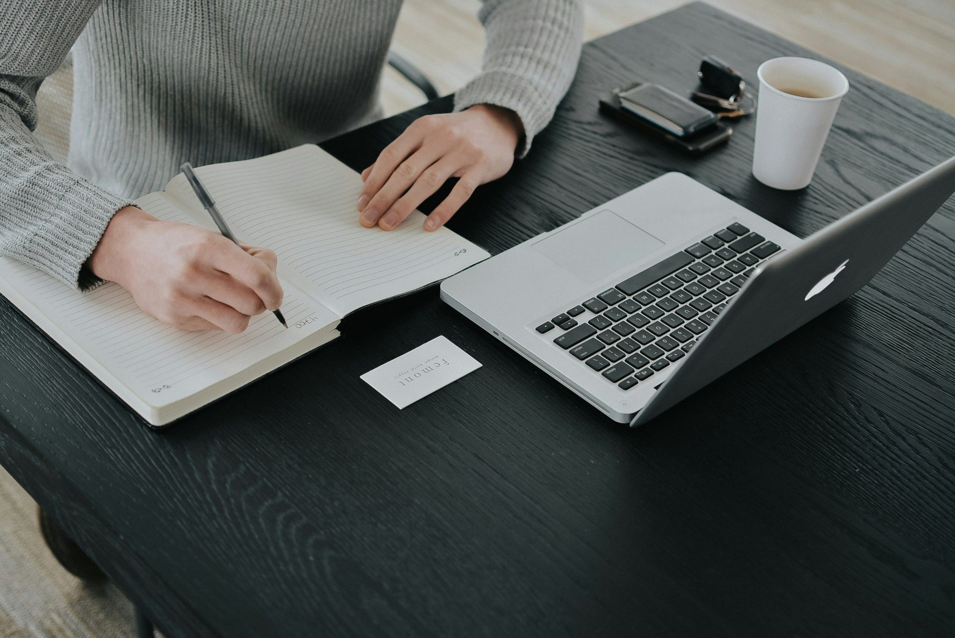 A person in a grey sweater writing in a notebook at a black desk